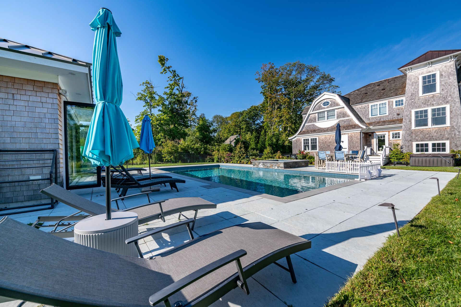 Poolside area with lounge chairs, blue umbrellas, and a house under a clear blue sky.