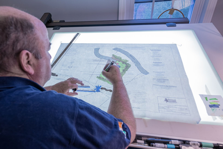 Man using a pen to draw on a lit architectural plan; studio setting.