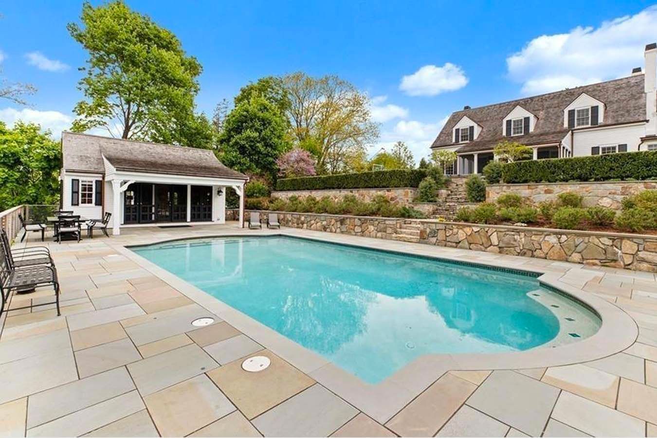Swimming pool with white house in the background and a pool house. Blue water, stone patio.